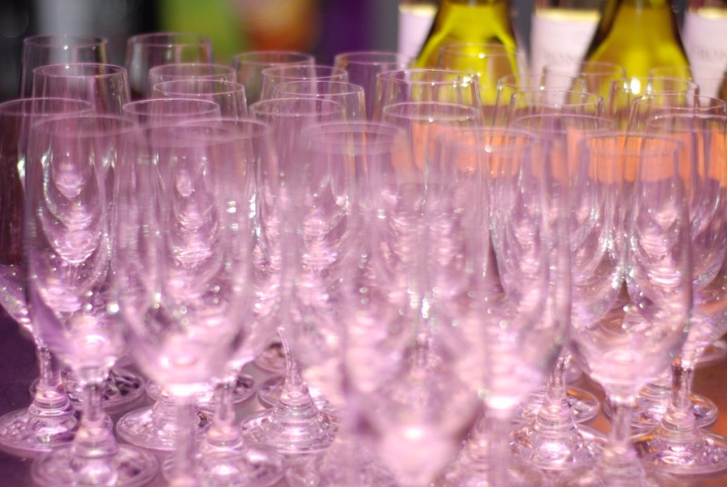 Rzędy pustych kieliszków do szampana ustawione na stole, z butelkami białego wina w tle, przygotowane na dużą uroczystość.